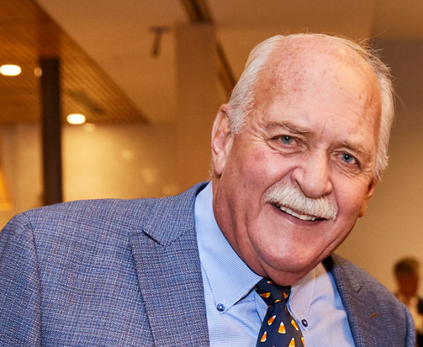 HEI Board Treasurer Jeff Lynch poses for a photo at an event. He has short, white hair, a white mustache, and wears a blue suit jacket and shirt.