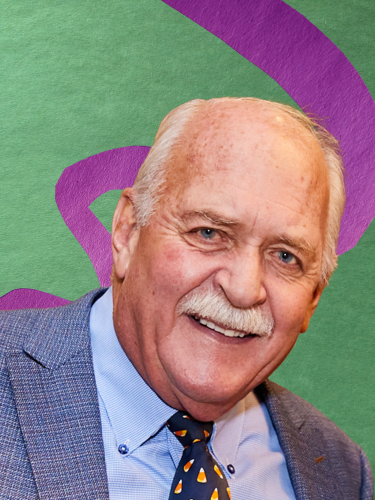 A white man with short white hair and a white mustache in front of a green construction paper background with a purple construction paper swirl. He is wearing a dark grey suit jacket, a light shirt and a dark tie. He is shown from the shoulders up.