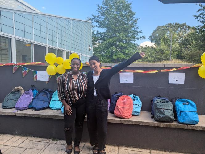 Two shelter workers pose in front of a row of backpacks at a back-to-school event.