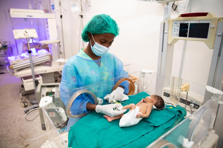 A NICU nurse treats a patient. The nurse wears a teal hair cover, a light blue medical mask, and a sheer blue medical gown over her flower-printed scrubs. The infant is in a plastic warming device that is open on the top. The baby wear a diaper and lays on a teal blanket. The baby is turned to the left side. The nurse is putting medicine into an IV on its foot.