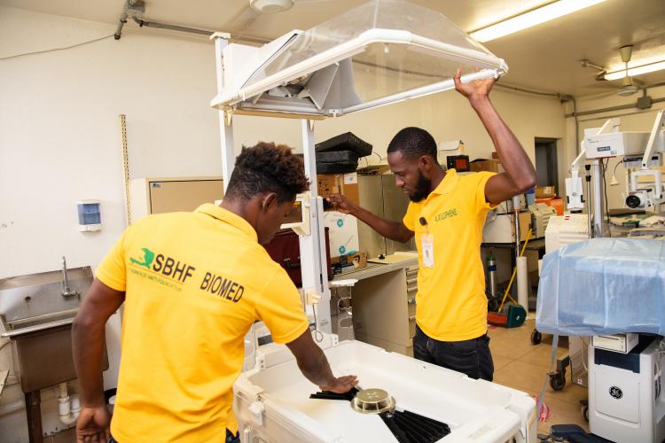 Two male Haitian biomedical repair technicians fix a neonatal incubator in a workshop.