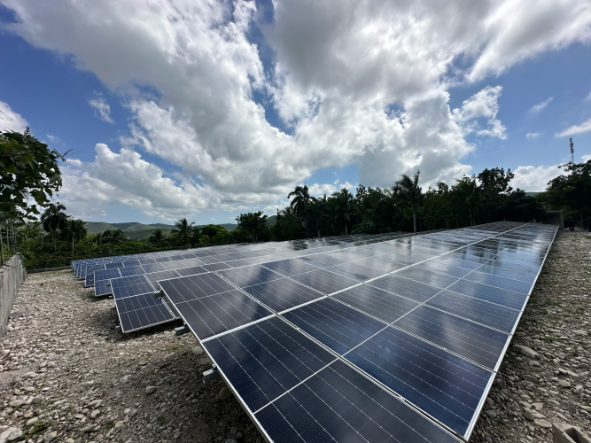 A solar panel field surrounded by tropical green trees and a bright blue sky with rolling clouds overhead.