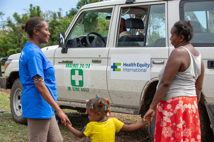 A young girl in yellow shirt and barrettes holds hands with Dr. Miliane Clermont, in blue T-shirt, and her aunt, who wears a red skirt. An HEI/SBH ambulance is behind.
