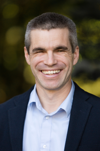 A headshot of Conor Shapiro smiling in a collared blue shirt and black jacket.