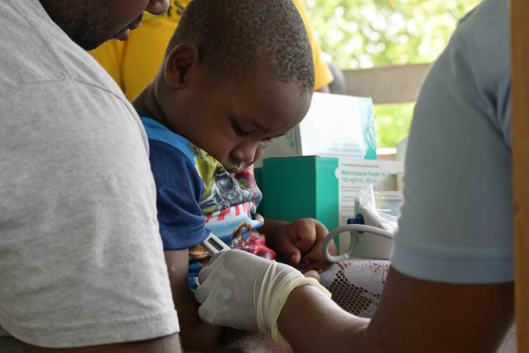A young child recieves medical care at a mobile clinic. The child is held by a man waering a white t-shirt. The child wears a colorful t-shirt and looks down at the nurse's gloved hand. The nurse's arm is visible on the right side of the screen.