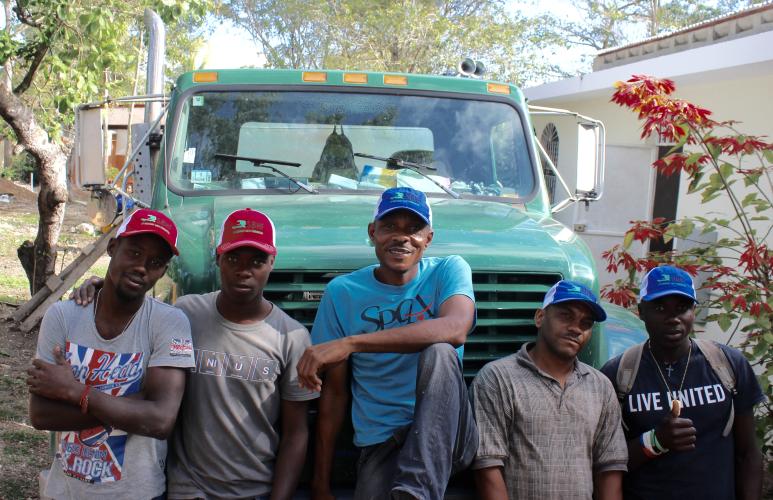 A group of five Haitian men sit in front of a large, light blue truck. They are all smiling and wearing blue or red baseball caps.