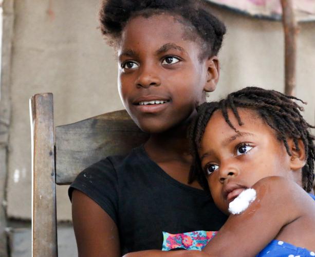 A young Haitian girl sits in a wooden chair and smiles while looking to her right. She has a younger girl in her lap. The younger girl in her lap who has a piece of cotton on her arm from a recent vaccination.