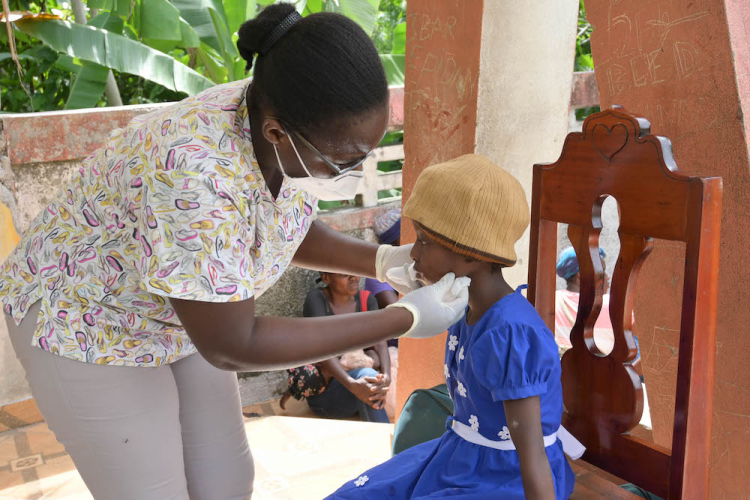 A nurse wearing scrubs leans over to put a bandage on a young girl's cheek. The girl sits in a wooden chair and wears a dark blue dress and a tan knitted cap.