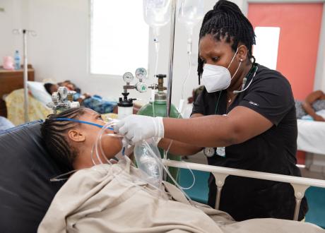 A woman patient lays in a hospital bed with a blanket covering her torso as a woman Haitian doctor puts an oxygen mask to her face. The doctor stands to the right of the bed. The doctor wears a white medical mask, black scrub top, and white exam gloves. Her hair is intricately braided. An IV pole stands to the left of the doctor, and to the left of the IV is a large green oxygen cylinder.