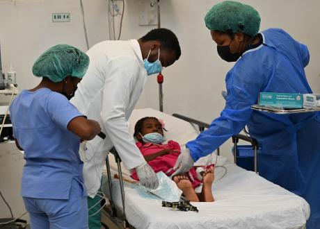 Three clinicians in surgical gear care for a young girl in pink dress and hair bow. She lies on a hospital bed with a metal device beside her.