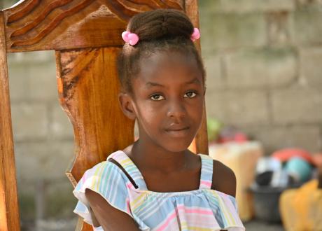 Head and shoulders of a 9-year-old Haitian girl smiling shyly at the camera. She wears a pastel dress and pink barrettes and sits on a large wooden chair.