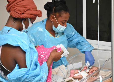 Two Haitian women look attentively at an infant in a hospital bassinet. The infant is waving bare limbs and his eyes and mouth are open. The women wear blue hospital gowns and face masks; one holds a bright pink onesie.