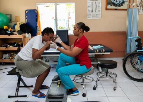 In a rehab center, two Haitian women sit across from each other, holding hands. Cherline leans forward, preparing to stand, while her therapist sits upright.