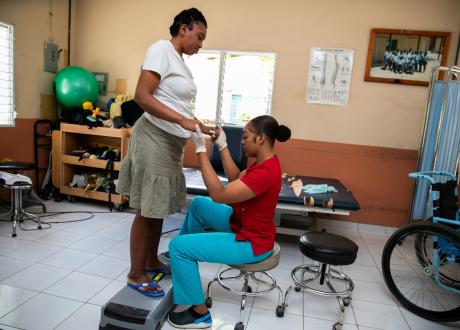In a rehab center, Cherline stands on a low step. Her therapist, in red and aqua scrubs, sits before her, holding her hands and looking down at her stance.