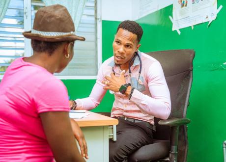 A Haitian male clinician sits behind a desk and speaks with a female patient, who sits with her back to the camera.