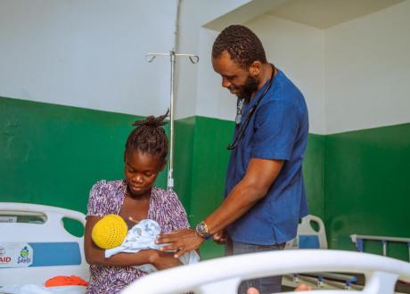 A male Haitian clinician consults with a woman who is holding her newborn baby. They are in a maternity ward at a hospital in Les Anglais, Haiti.