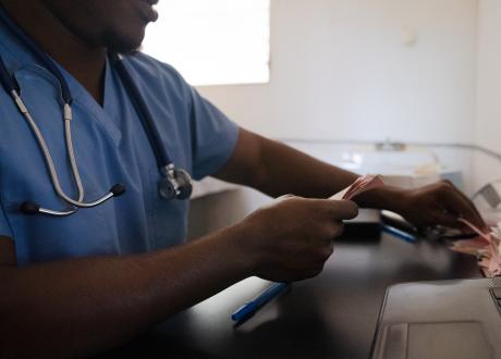A male Haitian doctor wearing light blue scrubs and a stethescope sits a a desk, shuffling through pink papers.