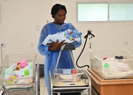 A Haitian woman stands behind her three newborn triplets, two of whom are in infant warming devices. Naromi holds the middle baby in her arms.