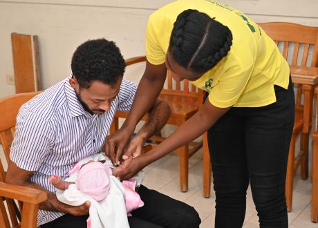 A light-skinned Haitian man is absorbed in the newborn he cradles on his lap, as a health worker in yellow polo shirt gives her a shot in the arm.