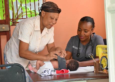 A Haitian woman leans over an examination table as she changes her baby. A Haitian woman doctor is sitting behind the table and is smiling at the baby.