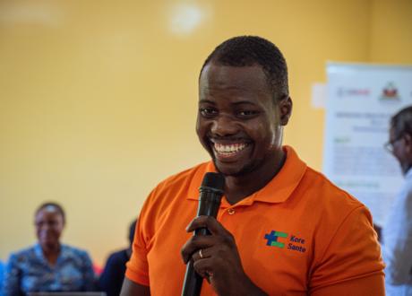 A Haitian man wearing a bright orange polo shirt holds a microphone and smiles brightly.