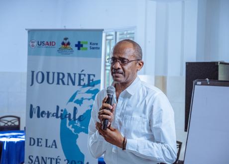 A Haitian male doctor speaks into a microphone. He has very short dark hair and wears a white button-down shirt. He holds a microphone with both hands close to his chest. Behind him is a display stand with the name of the conference on it.