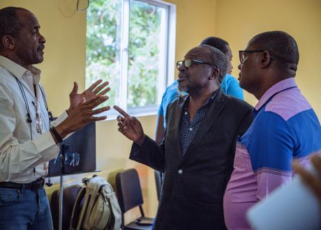 Three male Haitian doctors have a lively discussion in a yellow-painted room. They are gesturing to each other with their hands. 