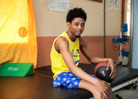 A young Haitian man named Galacxon sits on a physical therapy table and holds a ball in one hand. He is wearing a yellow basketball tank and blue and white patterned shorts. 