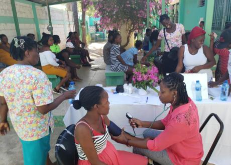 Pregnant Haitian women gathered in a covered outdoor space with bougainvillea blooming in the background. One is having her blood pressure taken.