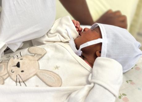 An infant rests peacefully on his mother’s lap. He is wearing a white cap and a cream-colored onesie with a rabbit appliqué.