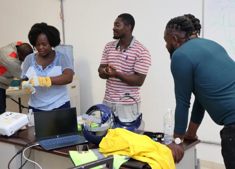 A staff member practices donning thick PPE gloves. Two other staff members observe.