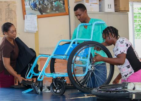 In a large, bright room, three Haitians wearing scrubs attach wheels to a wheelchair with a light-blue frame.