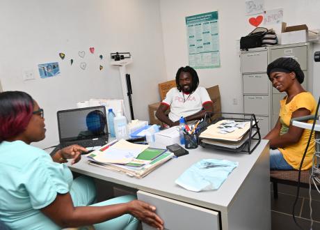 In a consultation room, a man with dreadlocks and a woman in a black head covering and yellow shirt smile as they talk with a woman in light turquoise scrubs.