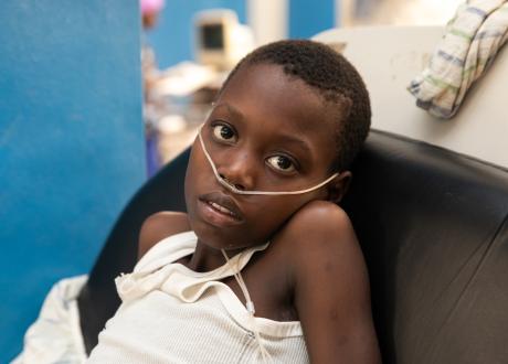 A young Haitian boy sits in an ER bed wearing a white tank top. He has a nasal cannula around the middle of his face.