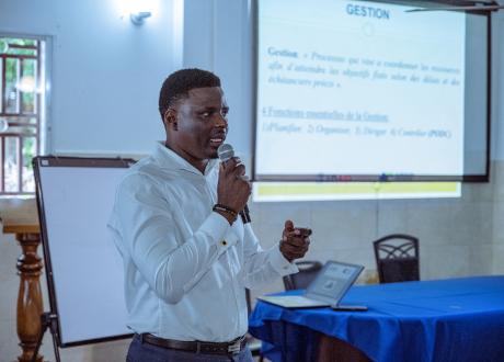 A Haitian male medical professional stands in front of a room and gives a presentation. He has short dark hair and wears a white button-down shirt. He holds a microphone and gestures with his free hand. Behind him, a projector displays one of his presentation slides.
