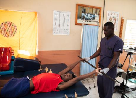 A Haitian man lies on a physical therapy table. He has his hands over his head and holds a long bar between them. A male Haitian physical therapist stands behind the table helping him with the exercise.