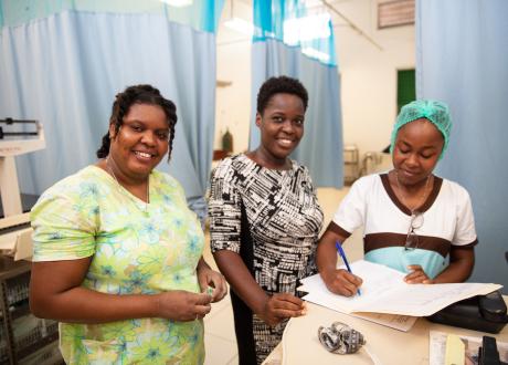 Three women Haitian maternal health clinicians stand by a desk and smile at the camera. 
