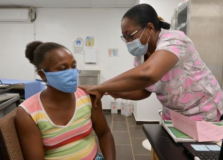 In a large exam room, one Haitian woman gives another a shot. Both wear face masks and have their hair pulled back. The nurse wears scrubs with pink hearts on them and the patient wears a brightly striped sleeveless top.