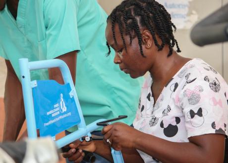 Close-up of a Haitian woman tightening a bolt on a section of wheelchair marked Free Wheelchair Mission. She wears scrubs, simple jewelry, and braids.