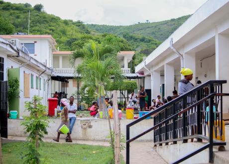 View of the hospital courtyard and green hills under a cloudy sky. The courtyard contains palm trees, many people, and colorful plastic containers of various sizes.