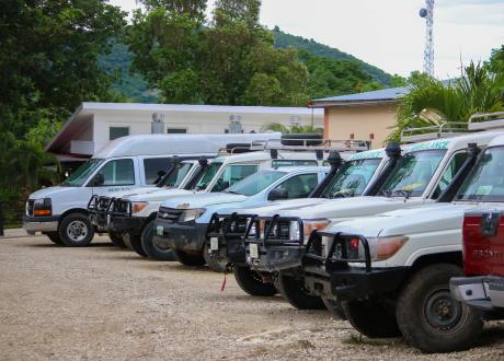 A half-dozen ambulances, a van, and a pickup truck parked in a row in front of hospital buildings, tree-covered hills behind.