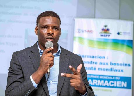 A Haitian man in a gray suit jacket and light blue shirt speaks into a microphone in front of a conference banner and a screen.