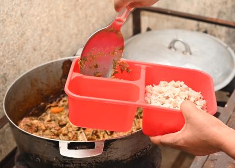 Close-up shot of hands spooning stewed mixed vegetables from a large aluminum pot into one compartment of a single-serving plastic container.