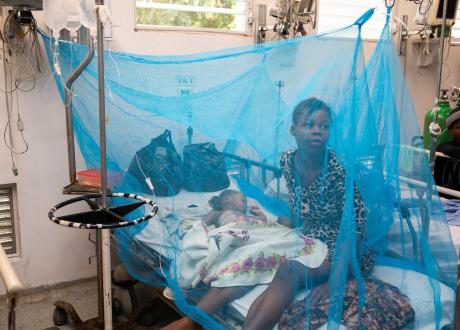 A young Haitian girl lays in an ER bed covered by light blue netting. Her mother sits besides her.