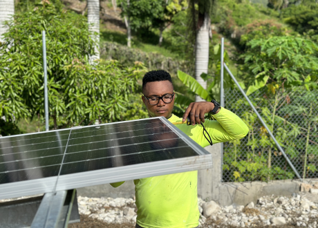 A Haitian technician screws a component into a solar panel in a lush green field.
