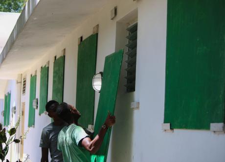 Two Haitian men position a piece of green-painted plywood over a glass window. The other windows of the sturdy white building are already covered by boards.