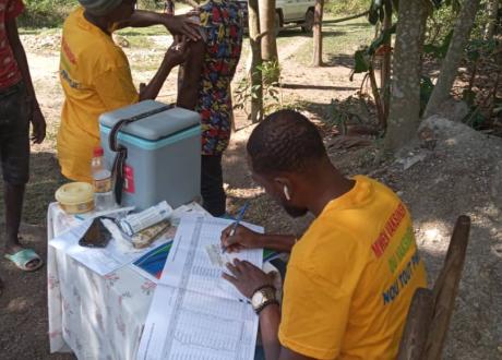 A community health worker sits at a small table outside and fills out paperwork. Behind the table, another community health worker administers a vaccine to a woman patient.