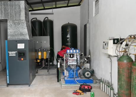 Interior view of a new cement building housing three large tall black tanks, ductwork, yellow- and blue-painted equipment, and green oxygen cylinders on hoses.