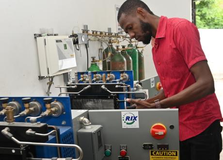 A slender Haitian man in a red shirt leans over a large piece of equipment with valves, filters, a control panel, and a caution sign.