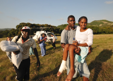 A Haitian man carries a woman across a field. The woman has his arm around his neck and has a bandage on her foot. A helicopter stands in the background.